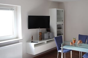 Ein modernes Wohnzimmer mit Flachbildfernseher, weißem Unterhaltungsmöbel, Glastisch und blauen Stühlen.