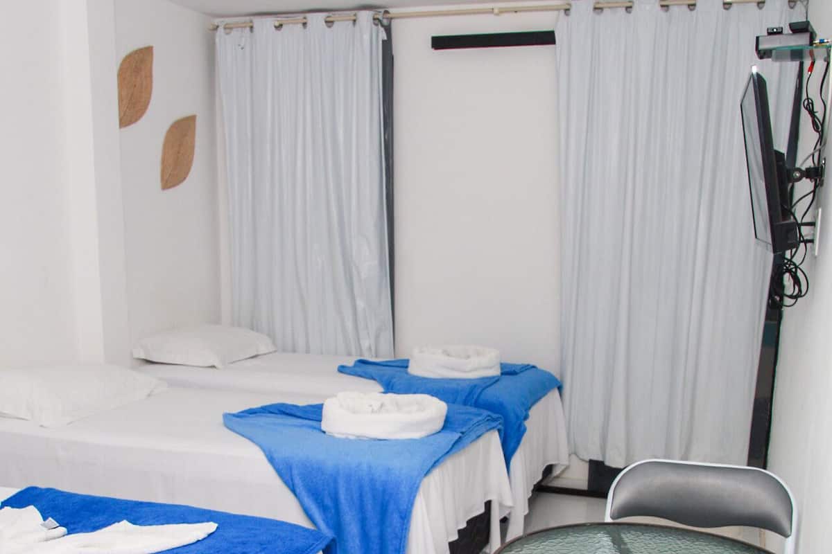 Habitación de hotel con dos camas, cada una con una toalla azul y ropa blanca. Hay una mesita redonda con una silla, y un televisor montado en la pared.