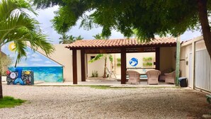 A covered outdoor seating area with wicker furniture, a mural of a beach scene, and a gravel ground. - Pousada Sereias de Maragogi (Maragogi)
