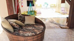 A wicker sofa with floral cushions in a covered outdoor area with a tiled floor and a view of a pool and garden. - Pousada Sereias de Maragogi (Maragogi)