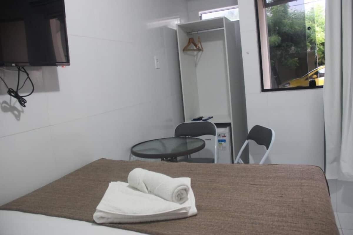 Habitación de hotel con cama, una mesita, una silla, un espejo y televisor montado en la pared.