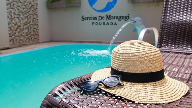 A straw hat and sunglasses on a wicker chair by a pool with a logo and name on the wall. - Pousada Sereias de Maragogi (Maragogi)