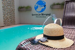 Un sombrero de paja y gafas de sol sobre una silla de mimbre junto a una piscina, con un logotipo y un nombre en la pared.