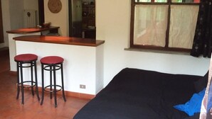 Ein Zimmer mit einem Bett, zwei Hocker, einer kleinen Bar und einem fenster mit Vorhängen.