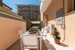 Ein Balkon mit Tisch und Stühlen, Blumentöpfen und Blick auf ein mehrgeschossiges Gebäude im Hintergrund.