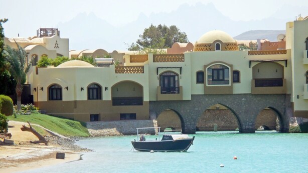 Ein Hotel an der Küste mit mediterraner Architektur, das gewölbte Fenster und eine Steinbrücke über dem Wasser zeigt.