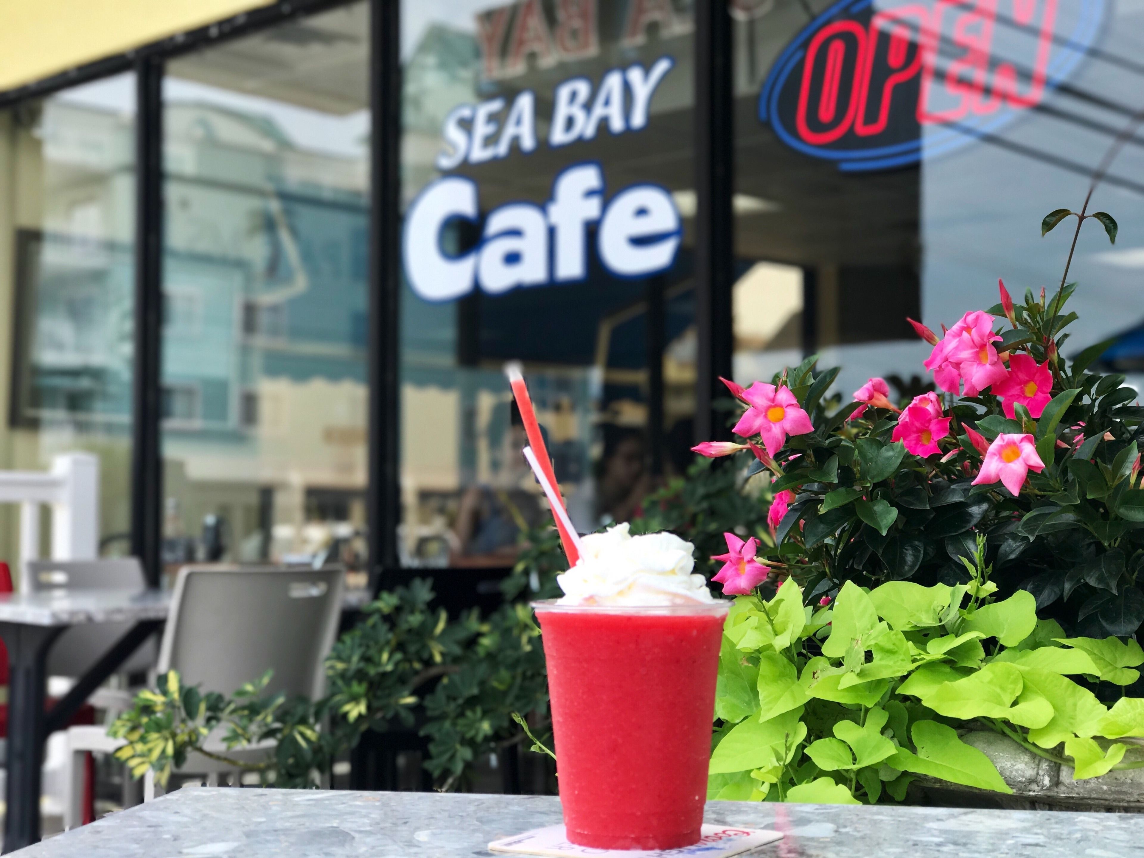 Punainen juoma, jossa on pilli, seisoo pöydällä kahvilan ulkopuolella. Kahvilan kyltissä lukee: ”SEA BAY Cafe”.