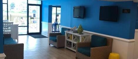 A hotel lobby with a blue wall, a flat-screen TV, and a seating area with a yellow ottoman.
