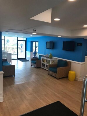 A hotel lobby with a blue wall, a flat-screen TV, and a seating area with a yellow ottoman. - Sea Bay Hotel (Ocean City)