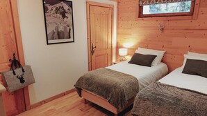 Une pièce aux murs lambrissés, avec deux lits, une table de chevet avec une lampe, un tableau encadré au mur et une porte en bois.