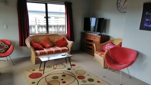 A living room with a wicker sofa, red chairs, a coffee table, a TV on a wooden cabinet, and a large window with curtains. - Caths cottage on Mayfair (Whangamata)