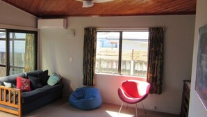 A room with a wooden ceiling, a ceiling fan, a dark sofa with cushions, a blue bean bag, a red chair, and a large window with curtains. - Caths cottage on Mayfair (Whangamata)