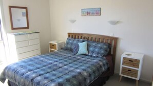 A bedroom with a bed, a nightstand, a dresser, and a wall-mounted shelf. - Caths cottage on Mayfair (Whangamata)