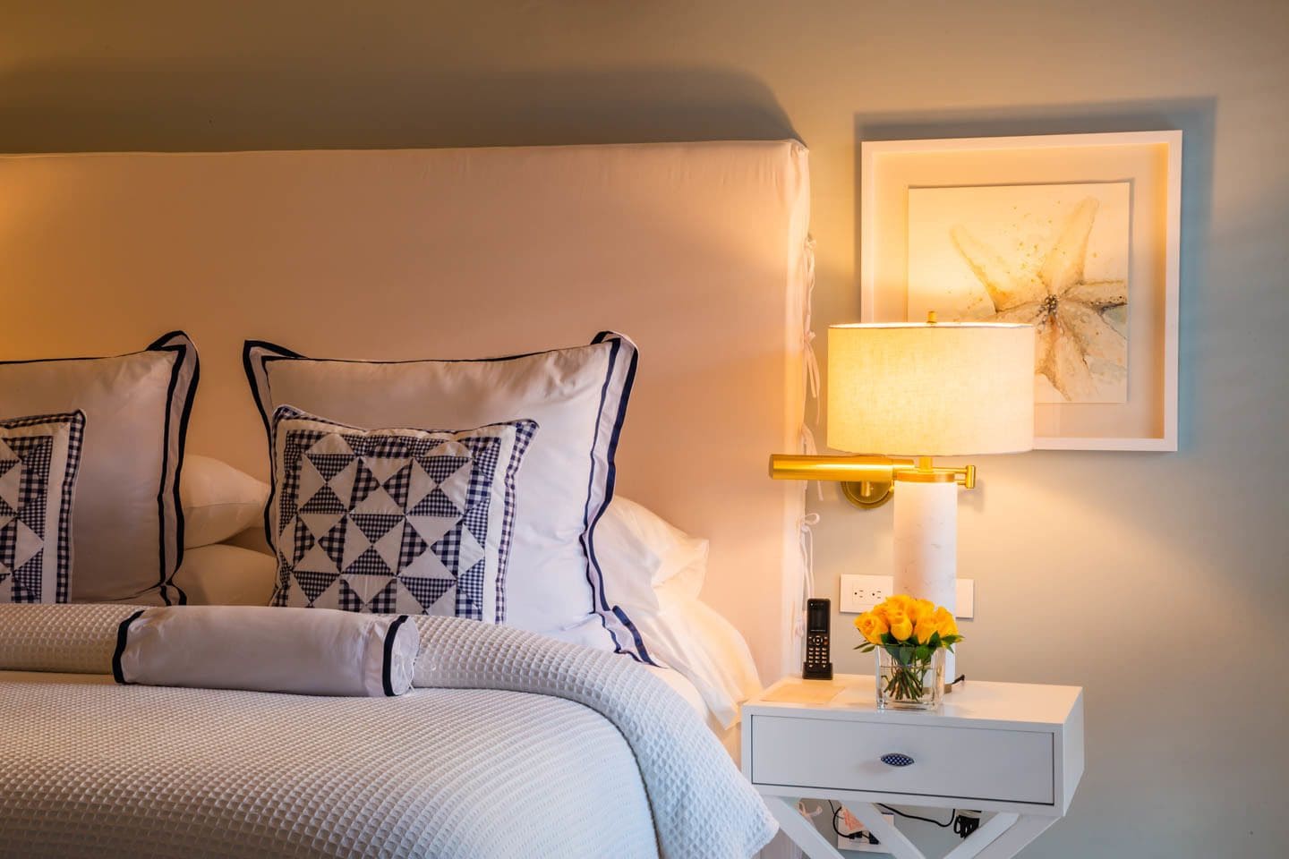 Una cama bien hecha con almohadones estampados, una mesita de noche con lámpara, un florero con flores y un cuadro en la pared.
