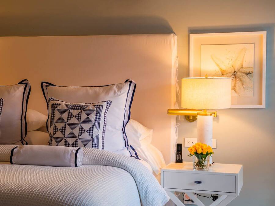 Una cama bien hecha con almohadones estampados, una mesita de noche con lámpara, un florero con flores y un cuadro en la pared.