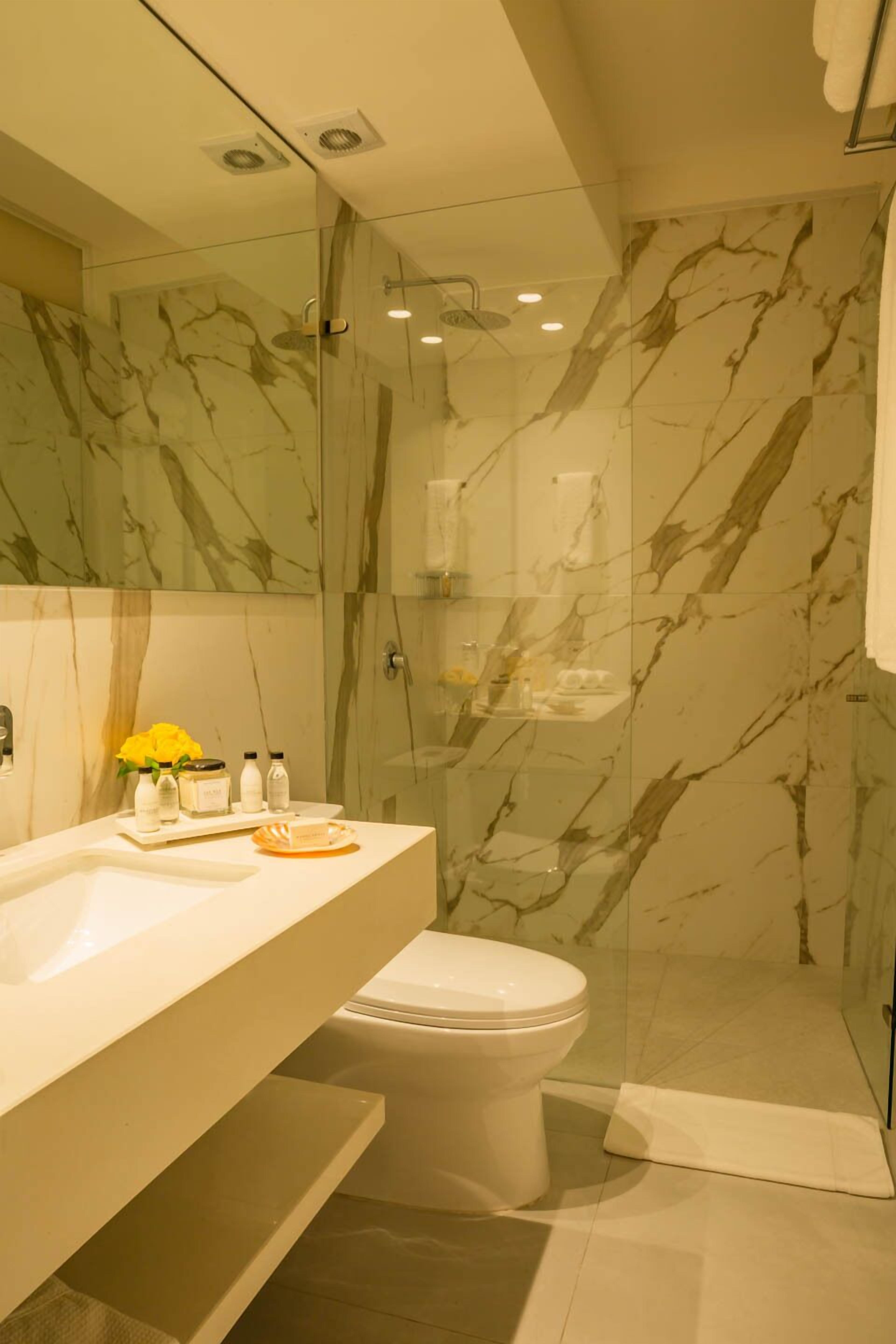 Un baño moderno con cabina de ducha de vidrio, pared con azulejos de mármol, lavamanos blanco y un inodoro.