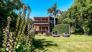 A modern two-story house with a wooden deck, surrounded by lush greenery and a clear blue sky. - Relax on Ridge | 7 seater car included (Oneroa)
