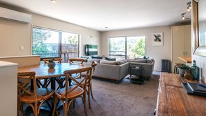 A spacious living area with a wooden dining table, comfortable seating, a grey sofa, and large windows offering a view of greenery. - Relax on Ridge | 7 seater car included (Oneroa)