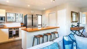 A modern kitchen with a central island, bar stools, and a dining area with a sofa and a mirror. - Relax on Ridge | 7 seater car included (Oneroa)