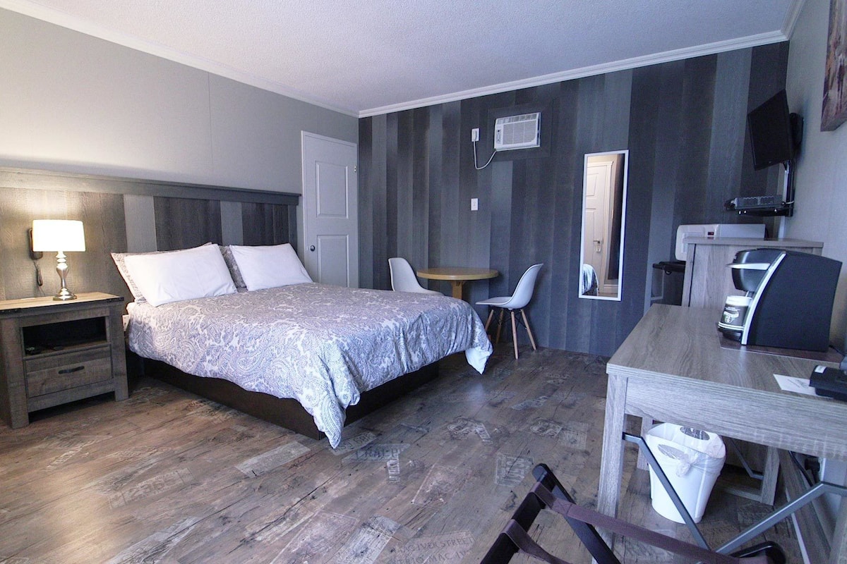 Une chambre d’hôtel comprenant un lit, une table de chevet avec une lampe, un bureau avec une chaise, une petite table ronde avec deux chaises, un miroir et une machine à café.
