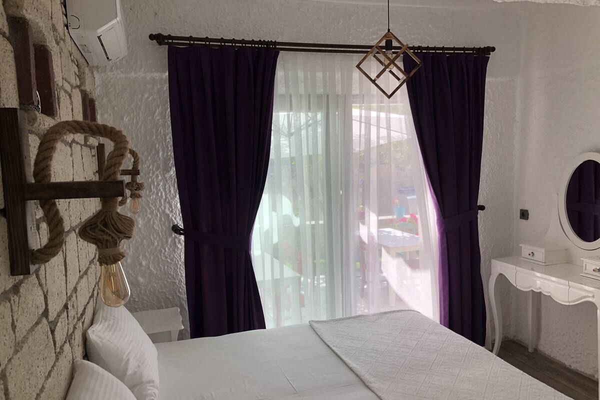 Un dormitorio con cama, una cruz de madera, una cuerda, una ventana con cortinas moradas, un tocador blanco y un espejo.