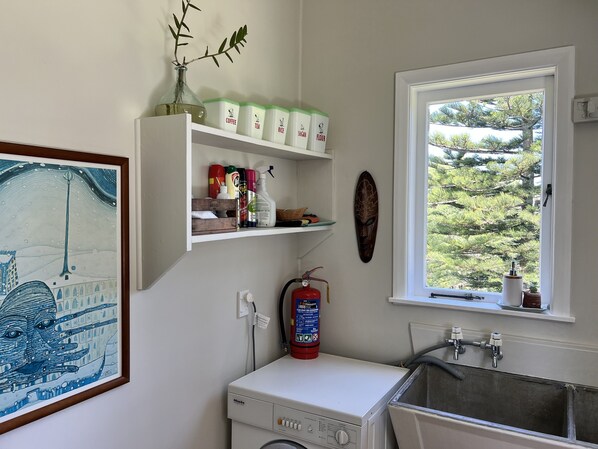 A laundry area with a washing machine, a fire extinguisher, and shelves with kitchen supplies. - Historic Farm Cottage with spectacular harbour/rural views (South Head (Kaipara))