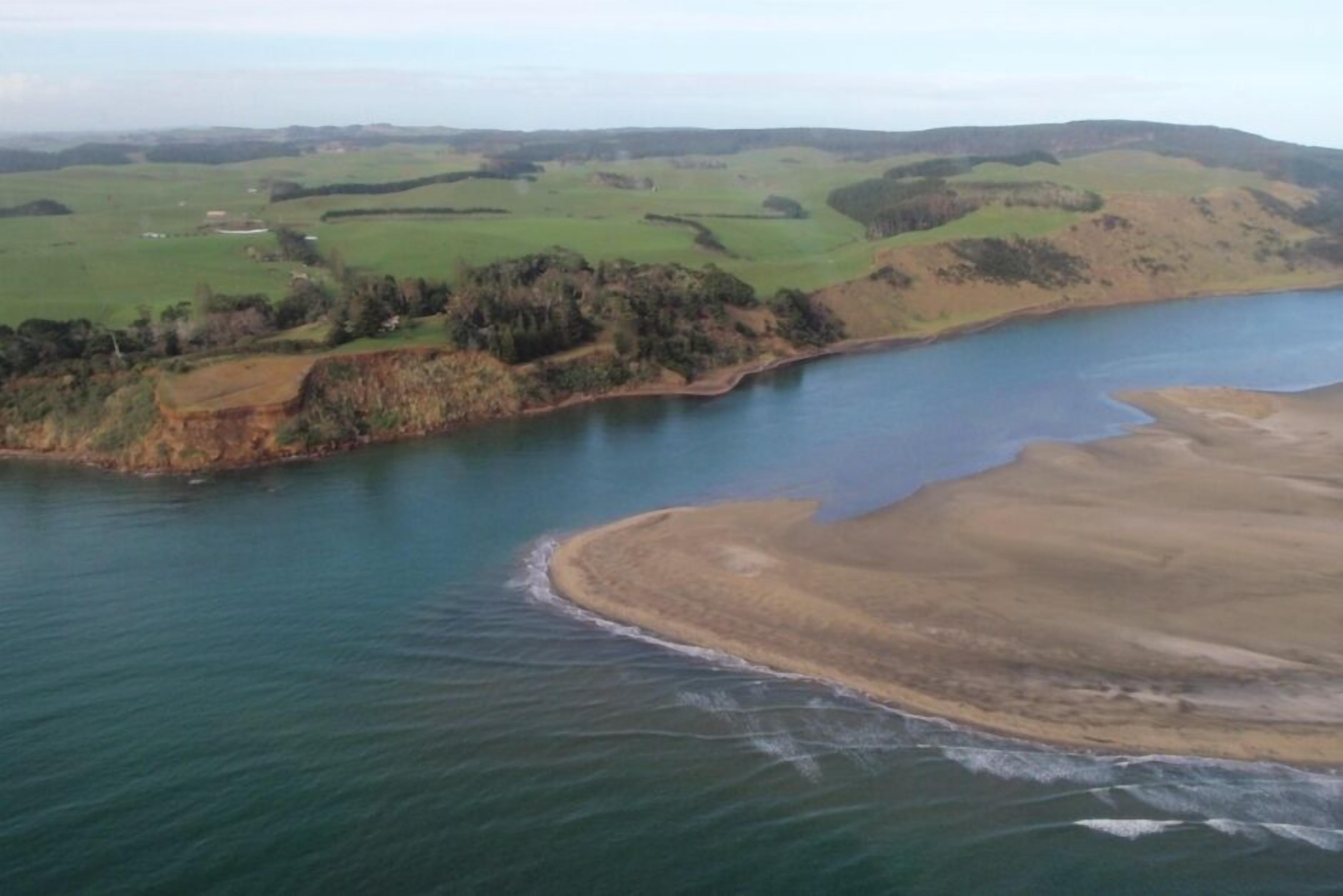 Vista aérea de un paisaje costero con una playa de arena, campos verdes y una masa de agua.