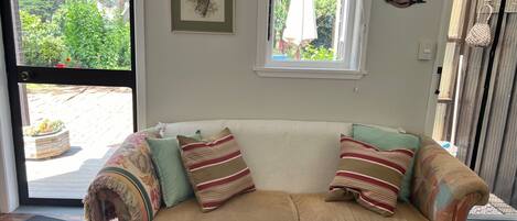 A cozy living room with a beige sofa, patterned cushions, a framed artwork, and a window with a view of greenery.