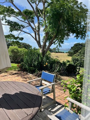 A wooden deck with a blue chair and a table, surrounded by lush greenery and a view of the sea. - Historic Farm Cottage with spectacular harbour/rural views (South Head (Kaipara))