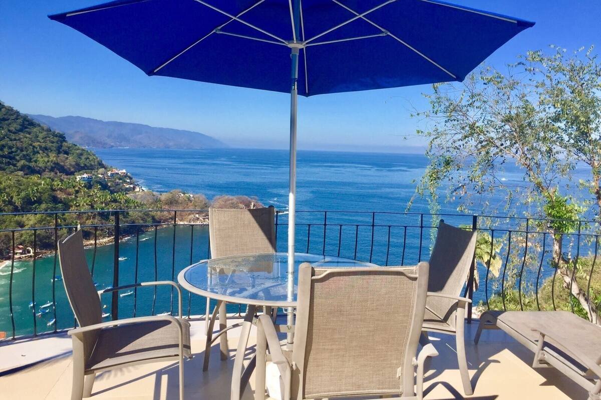 Ein Balkon mit Glastisch und Stühlen, einem blauen Sonnenschirm und Blick auf das Meer und die Hügel.