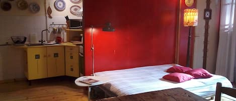 A compact room with a bed, a small table, a wooden desk, a kitchenette, and a red accent wall.