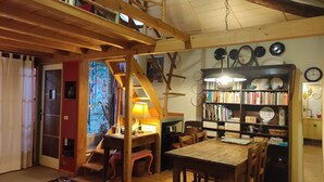 A cozy, rustic room with a wooden staircase, a bookshelf filled with books and decorative plates, a dining table with chairs, and a patterned rug on the floor. - Baubaus House_Holidays house in Cuirone (Vergiate)