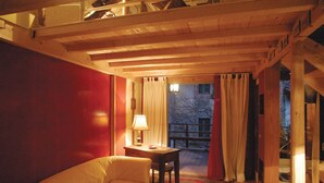 A cozy room with a wooden ceiling, a red wall, a beige sofa, a small table with a lamp, and a view through a window with curtains. - Baubaus House_Holidays house in Cuirone (Vergiate)