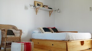 A bedroom with a wooden bed, a wicker chair, a small bedside table, and a shelf with books and a picture frame.