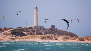 A lighthouse on a hill overlooking the sea with several kites flying above.