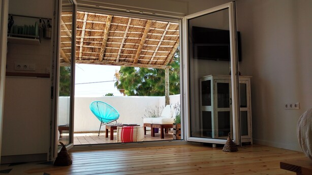 A room with a sliding glass door leading to an outdoor area with a thatched roof, a blue chair, a white sofa, and a cabinet.