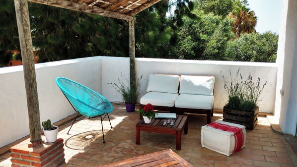 A covered patio with a wooden pergola, a turquoise chair, a white sofa, a coffee table, and potted plants.