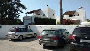 A parking area with three cars parked in front of a building with white walls and a thatched roof.