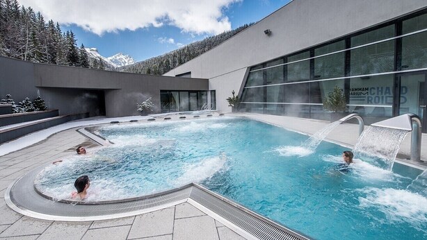 Ein moderner Poolbereich im Freien mit Whirlpool, umgeben von einem Gebäude mit großen Fenstern und Blick auf schneebedeckte Berge.