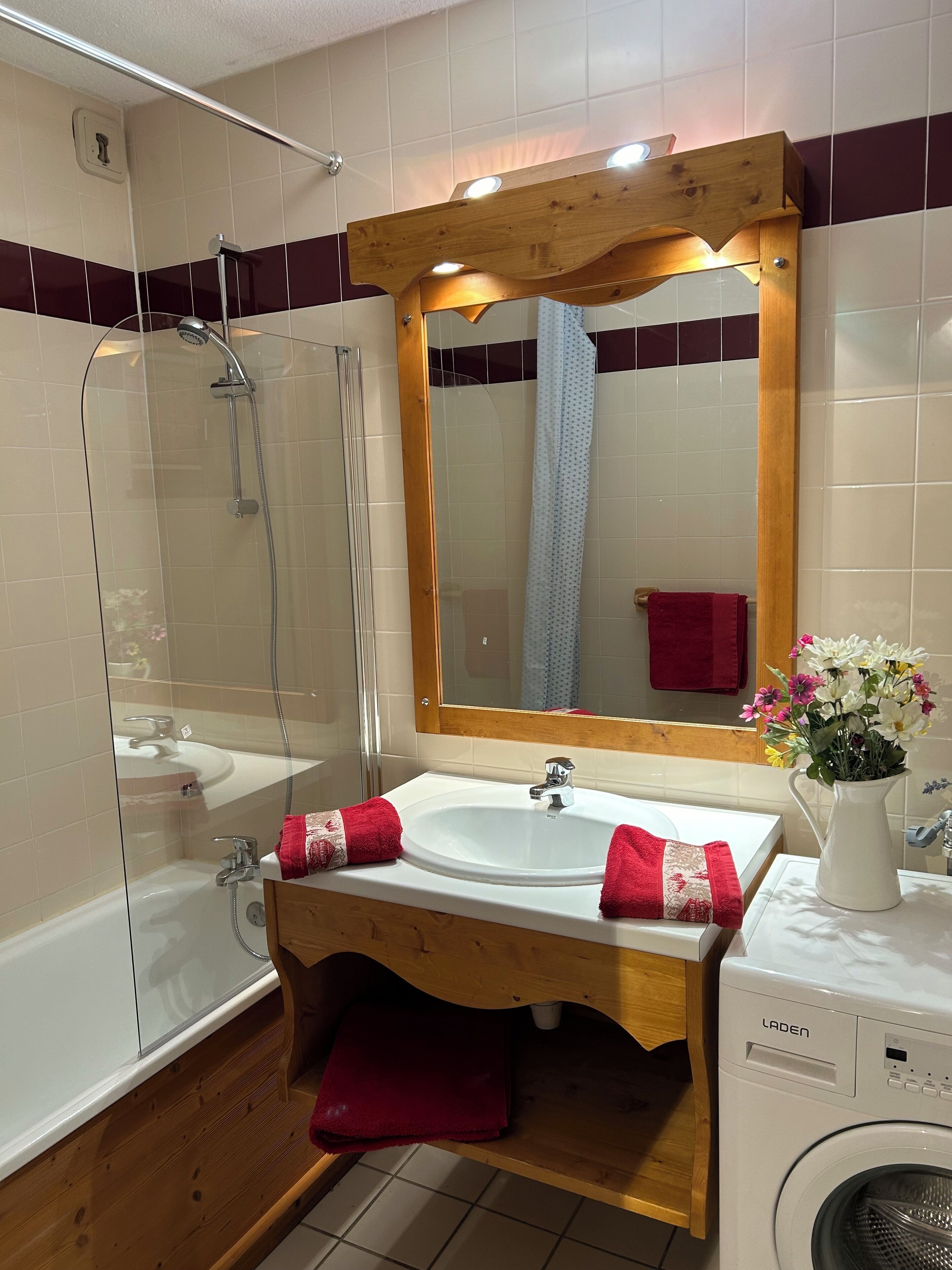 Un baño con ducha de vidrio, un lavabo blanco con tocador de madera, un espejo con marco de madera y una toalla roja sobre la encimera.
