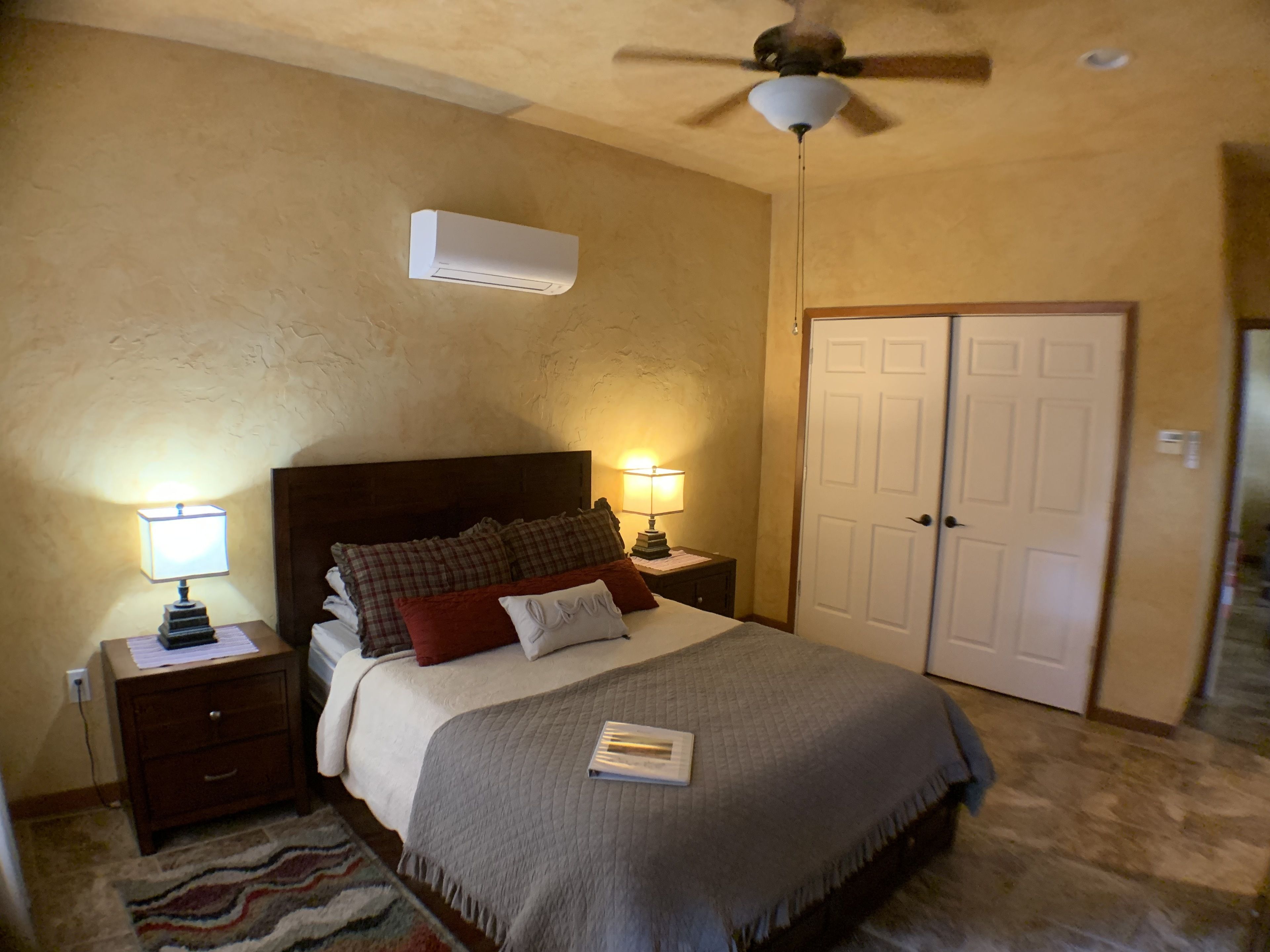 A bedroom with a bed, two bedside tables with lamps, a ceiling fan, an air conditioning unit, and a door.