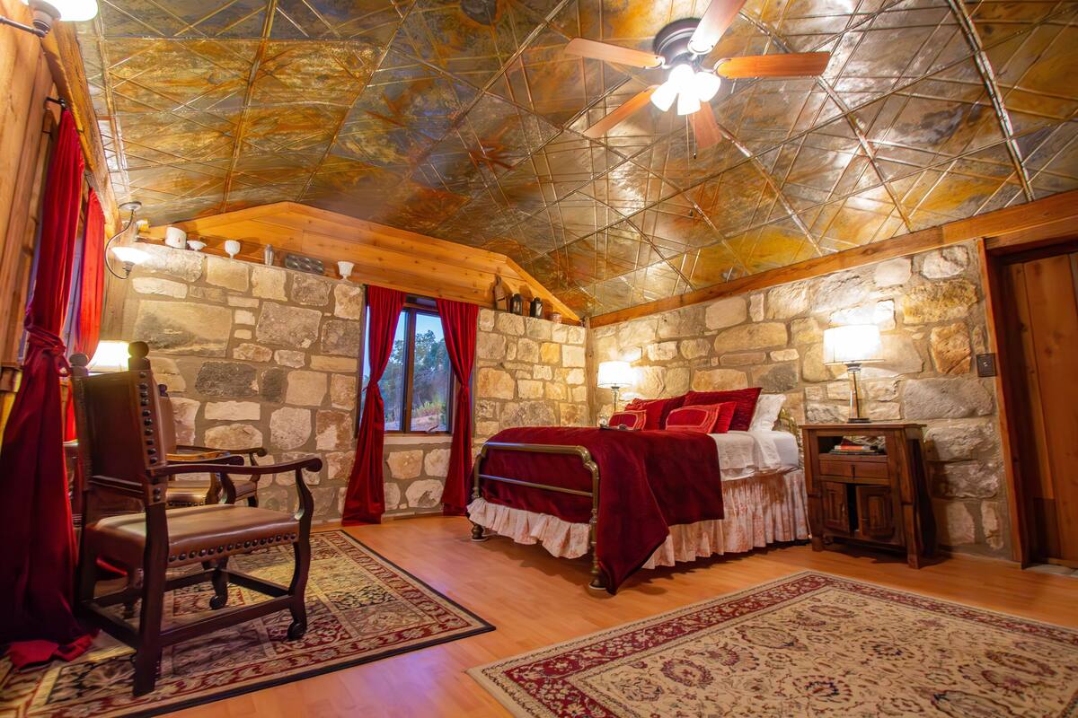A cozy bedroom with a stone wall, a wooden ceiling, a bed with red bedding, a chair, and a nightstand with a lamp.