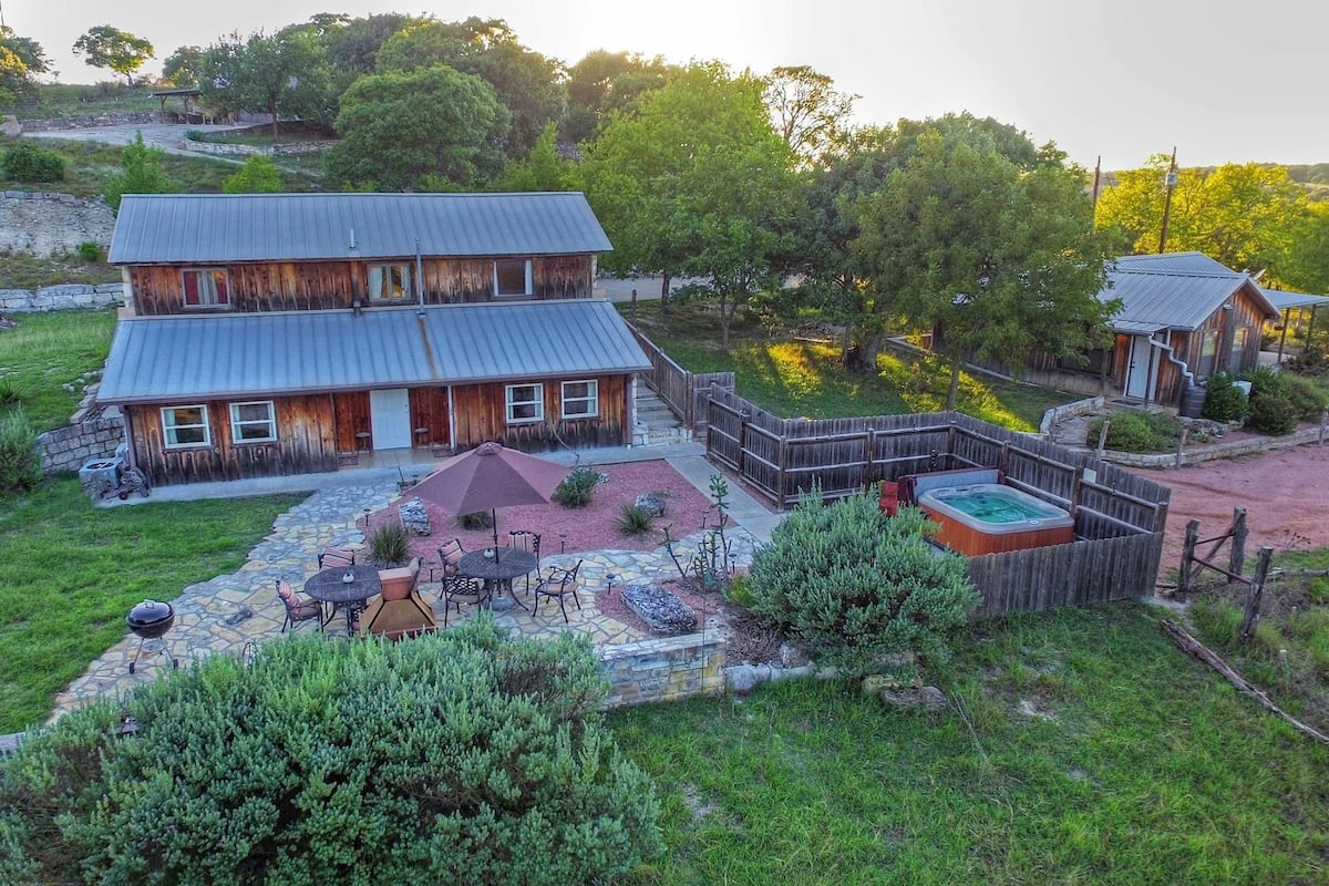 A rustic two-story wooden house with a stone patio, outdoor seating, and a hot tub.