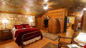A cozy log cabin-style room with a stone wall, a wooden bed with red bedding, a wooden door, and a ceiling fan. - A Barn at the Quarry (Fredericksburg)