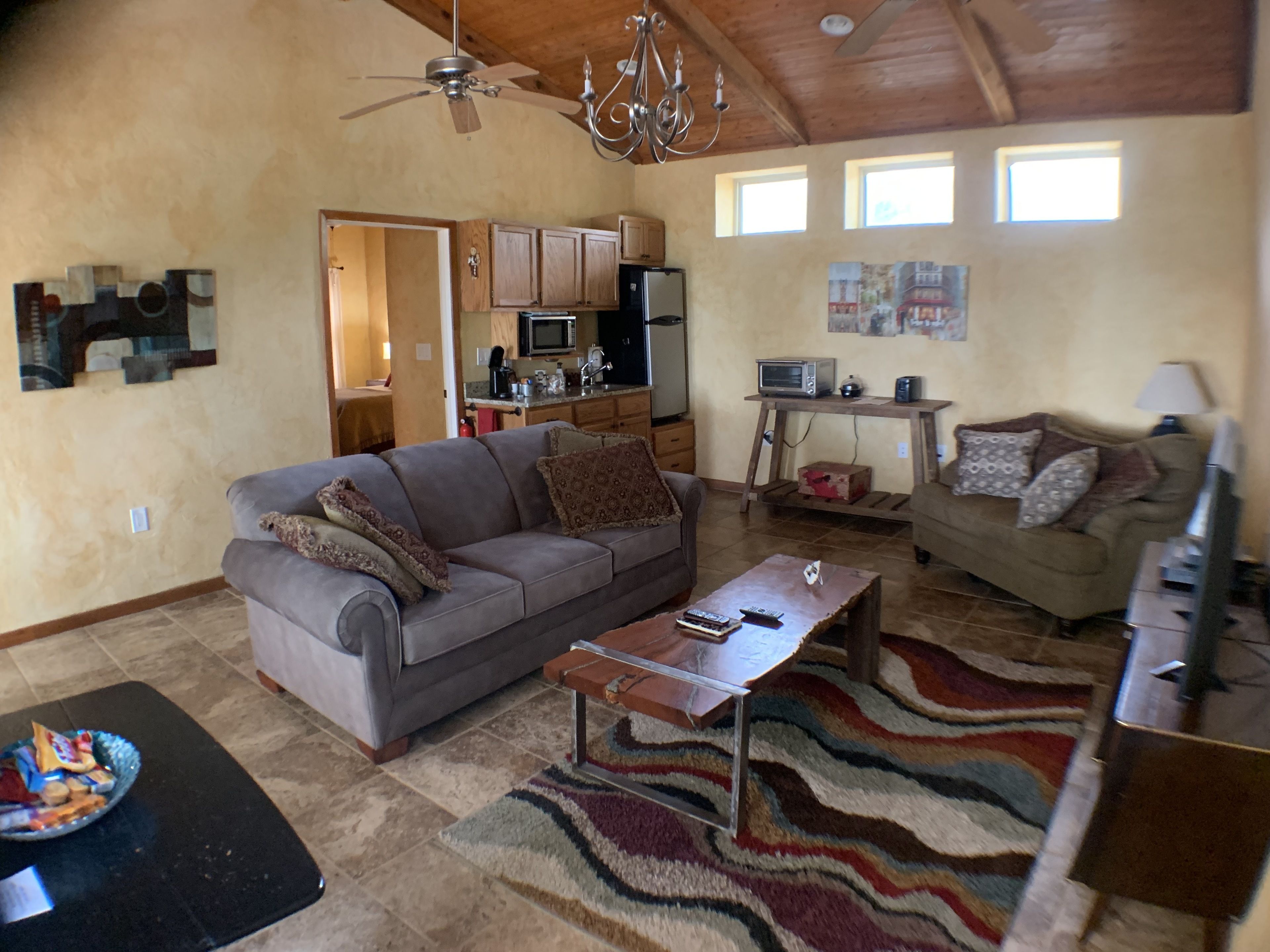 A spacious living room with a grey sofa, a coffee table, and a patterned rug. A kitchen with wooden cabinets and a microwave is visible in the background.