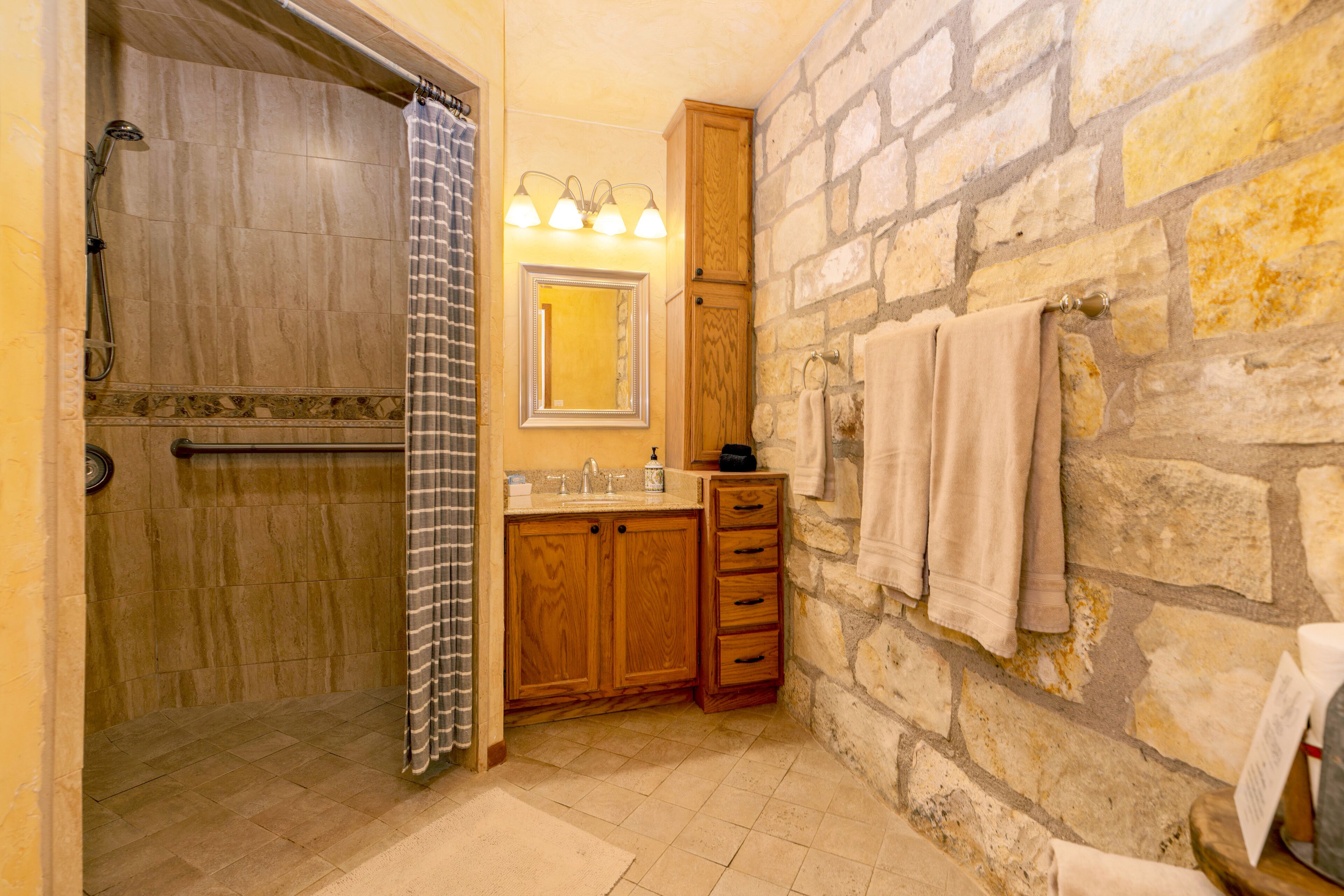 Un baño con ducha, lavamanos, armarios de madera, espejo y paredes de piedra.