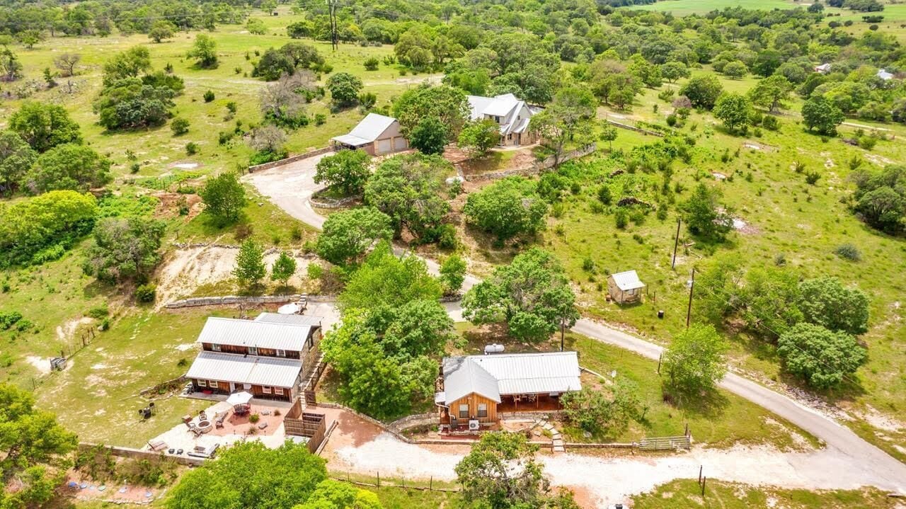 Il s’agit d’une propriété rurale comprenant deux bâtiments principaux : une grande maison dotée d’un patio et un petit chalet, le tout entouré d’une végétation luxuriante et bordé d’un chemin de terre.
