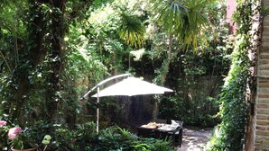 A lush garden with a table and chairs under a white umbrella, surrounded by dense foliage and potted plants.
