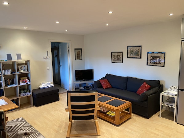 A living room with a sofa, TV, bookshelf, and a small table with a chair.