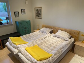 Una cama bien hecha con ropa de cama escocesa y dos almohadones amarillos, una mesita de noche con una lámpara, una ventana con vistas a la vegetación y un mueble verde.